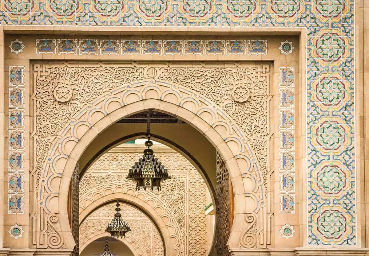 The arches outside the Royal Palace in Casablanca, with intricate yellow designs and blue tiles. Lights are hanging from each archway as you look through.
