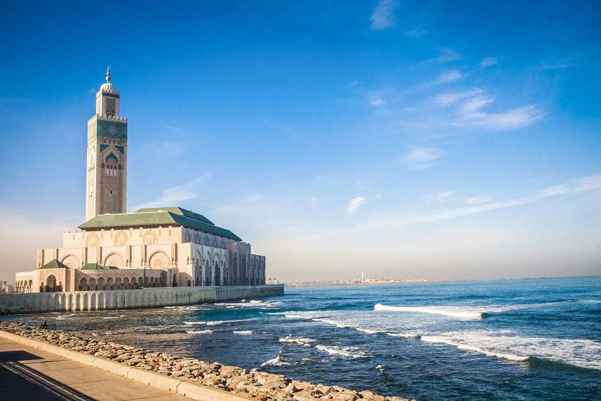 A view of the Hassan II Mosque in Casablanca Morocco with the ocean and soft, rolling waves in the foreground on a clear evening. 
