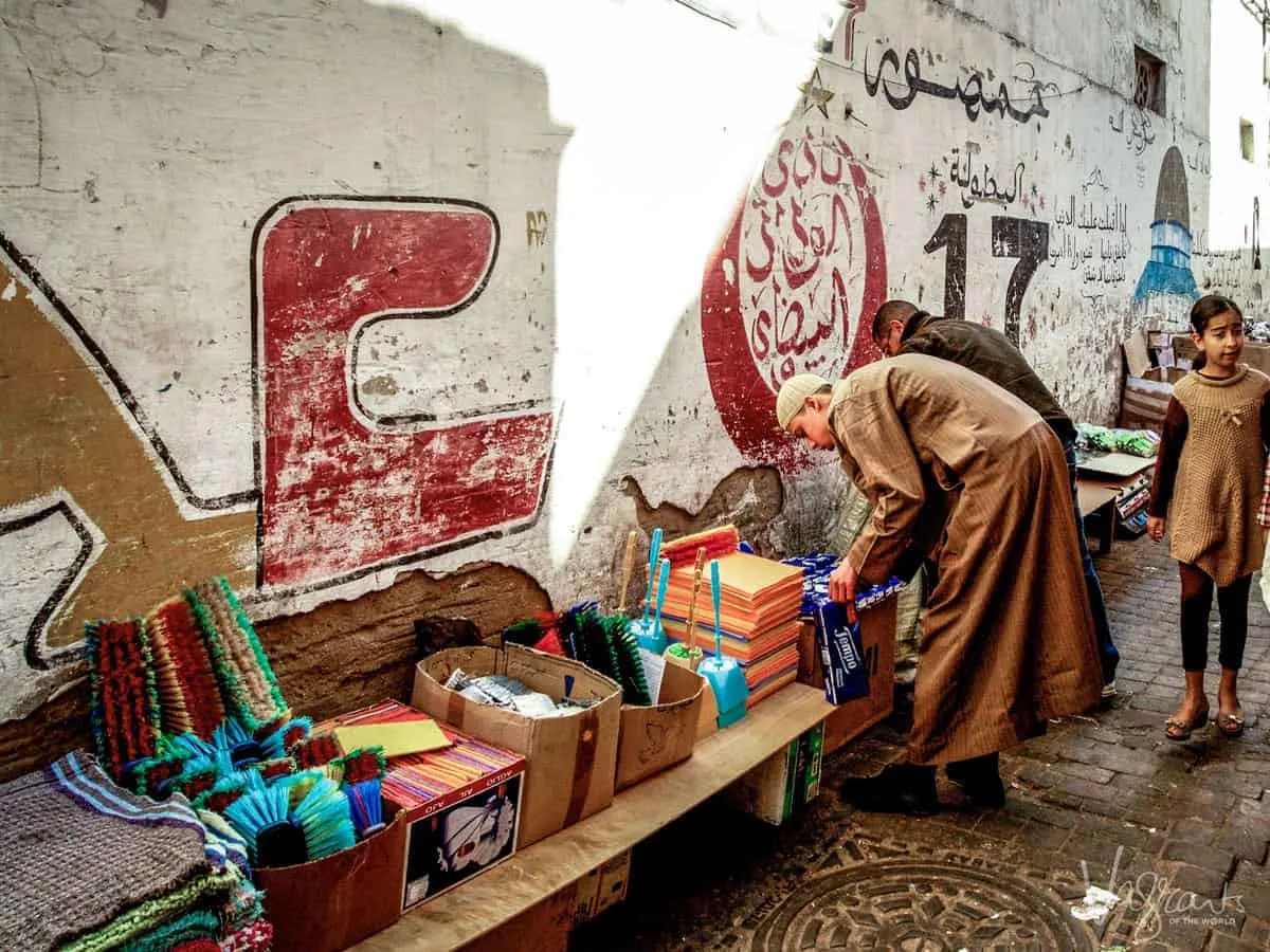 Two men bent over looking at products on a bench at a market in Casablanca, with a young girl in the edge of the photo looking away. 