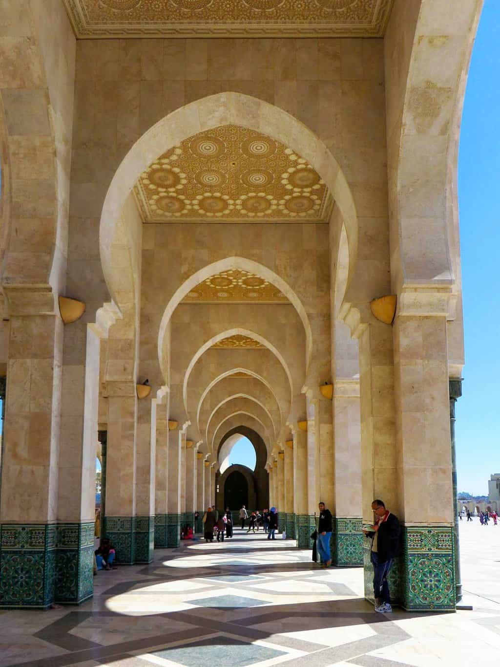 An arched covered walkway with intricate designs covering a patterned marble floor outside the Mosque in Casablanca. 