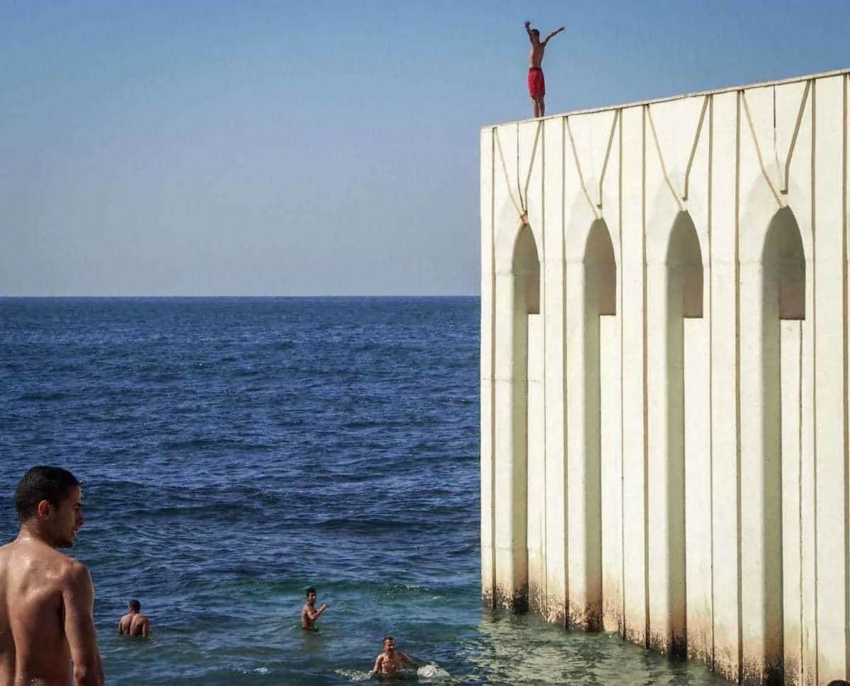 A young man about to dive into the ocean from atop a white wall. Four other young men are already in the water. 
