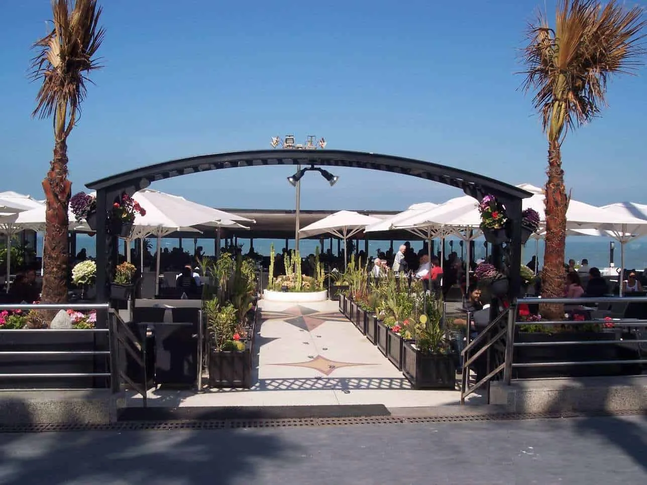 A walkway under an arch with restaurant tables with umbrellas on both sides in Corniche, Casablanca, Morocco.
