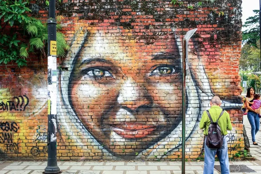 A man in a green shirt standing in front of a mural of a smiling girl with dark skin. 