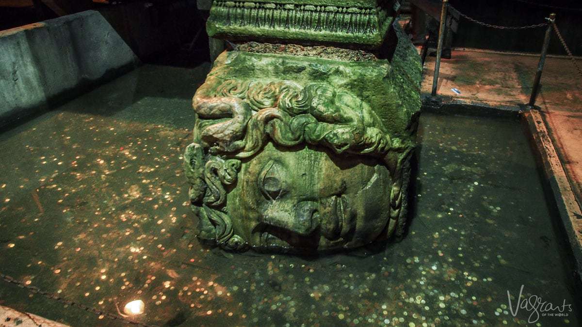 Medusa Head carved in the base of a pillar placed in water surrounded by coins. 