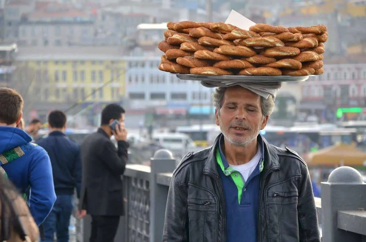 A man carrying tray of bagels on a tray on his head through the streets. 