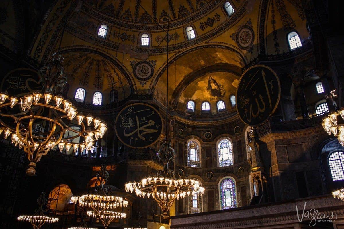 The domed ceiling of the Hagia Sophia in Istanbul with golden frescoes and stain glass windows. Hanging chandeliers provide light in the dimly lit room. 