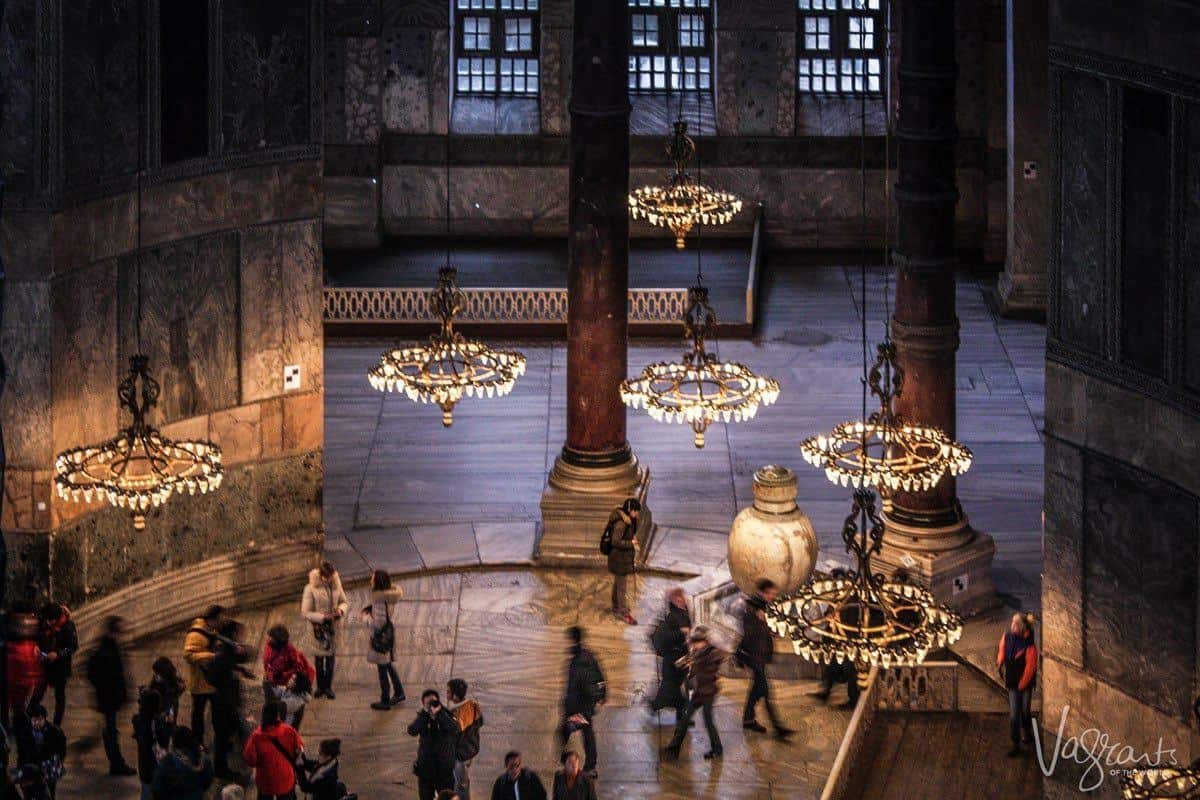 Tourists inside the Hagia Sophia mosque in Istanbul. The light is low and circular chandeliers of lights hang around the large room. 