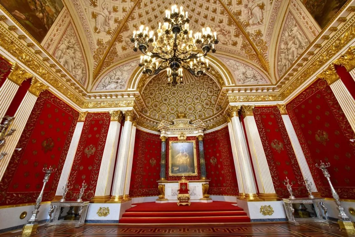 A royal looking room with red walls and golden ceiling lit by a massive chandelier in the Hermitage, St Petersburg Russia. 