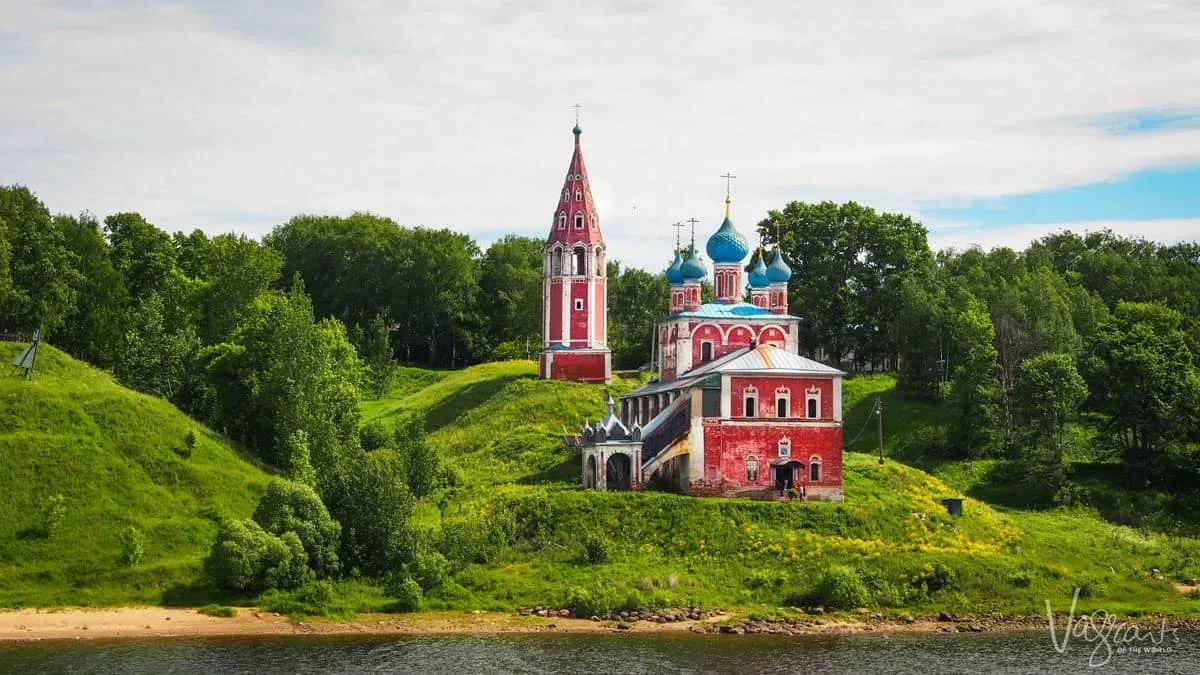 red and blue church set on emerald green grass on the banks of a river during a cruise from st petersburg to moscow.