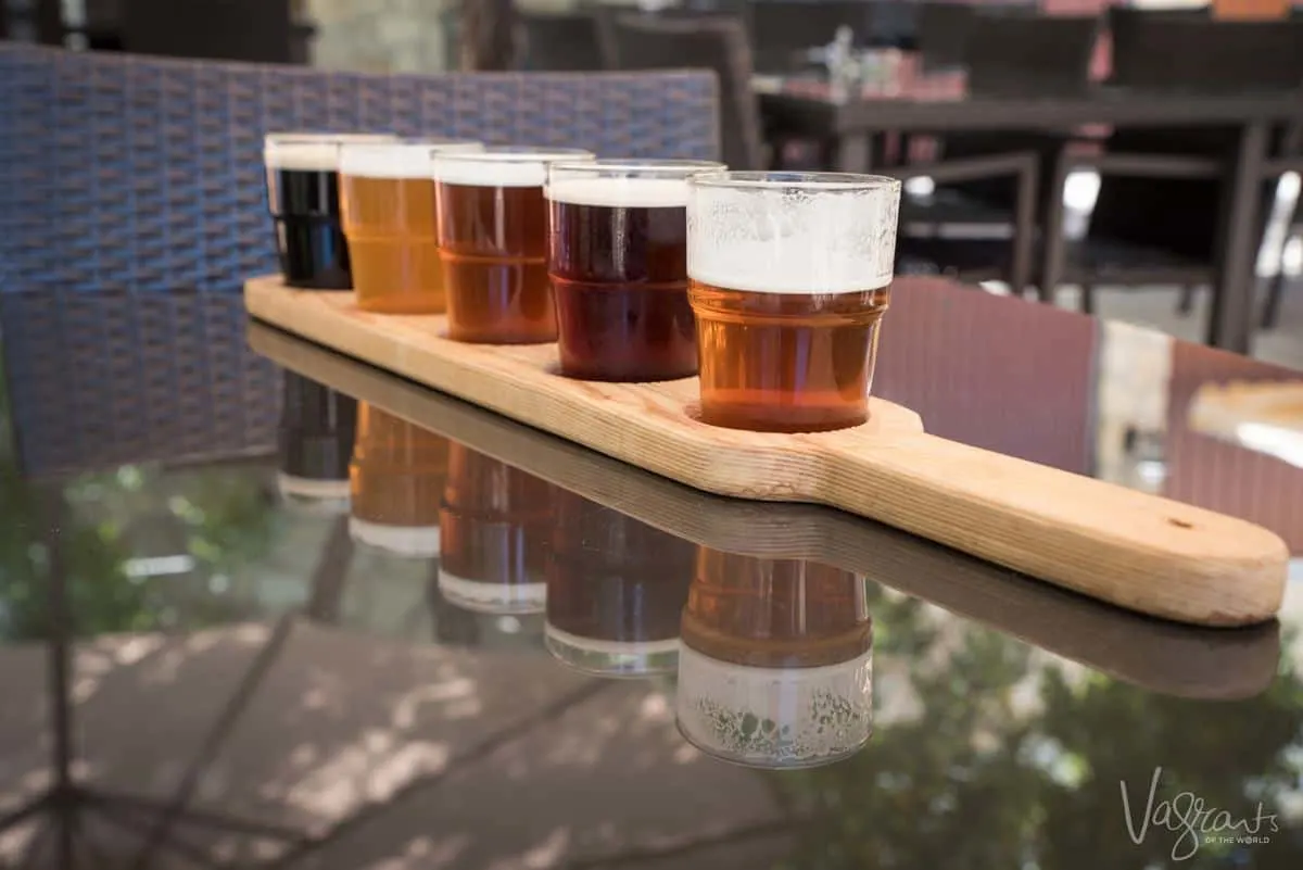 A row of different coloured beers in a wooden beer flight board reflected in the glass table. 