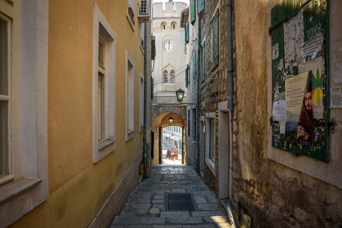 Poeple walking through an archway with a clocktower above at the end of a narrow lane in Herceg Novi Old Town in Montenegro. 