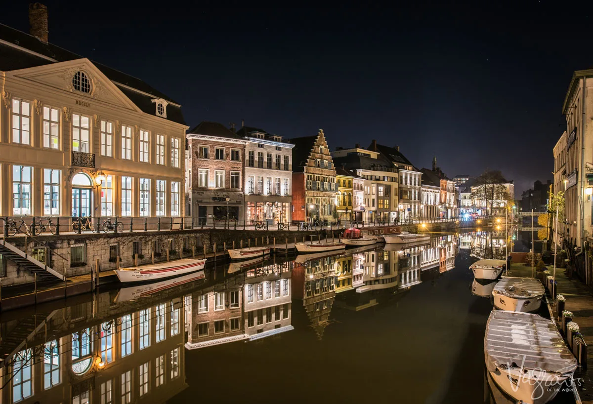 Colourful houses lit up at night reflect in the water on the canals of Ghent where small canal boats are tied up along side.