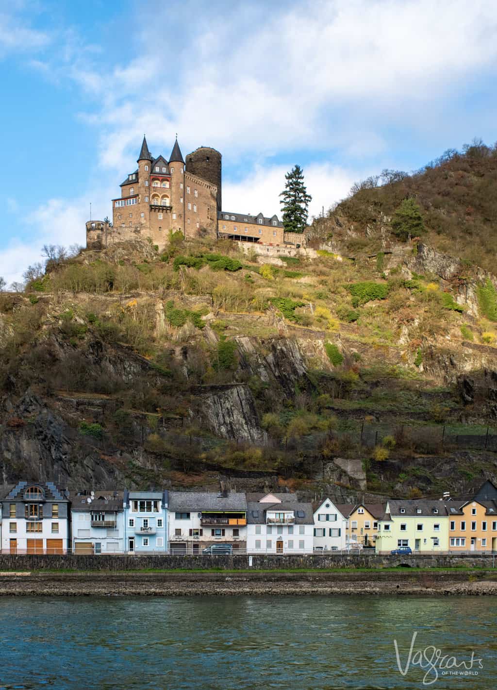 A castle perched on top of a rocky outcrop above a small village on a river bank in Germany. 