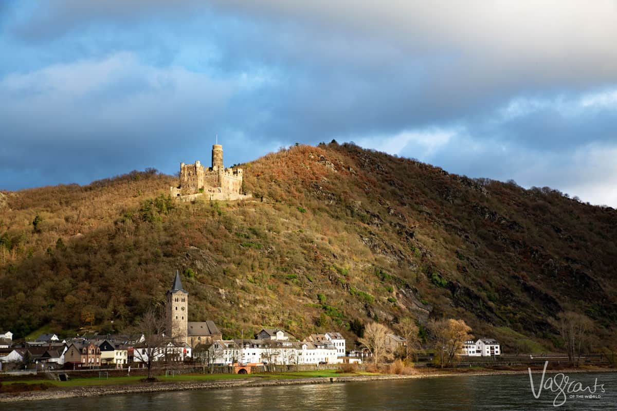 A castle sits on a hill above a small traditional village on the Rhine river.