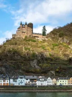 A castle perched on top of a rocky outcrop above a small village on a river bank in Germany.