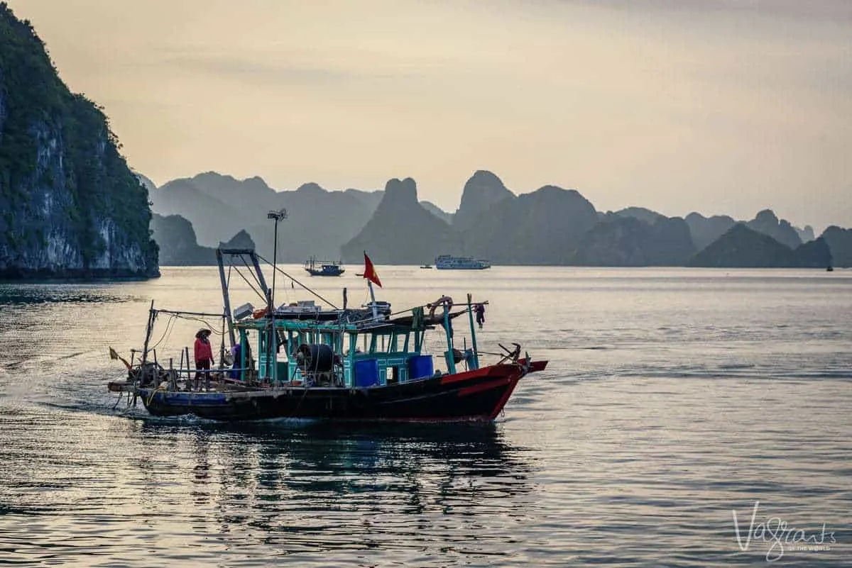 A lady in a traditional Vietnamese hat stands on a brightly coloured fishing boat at sunrise. 