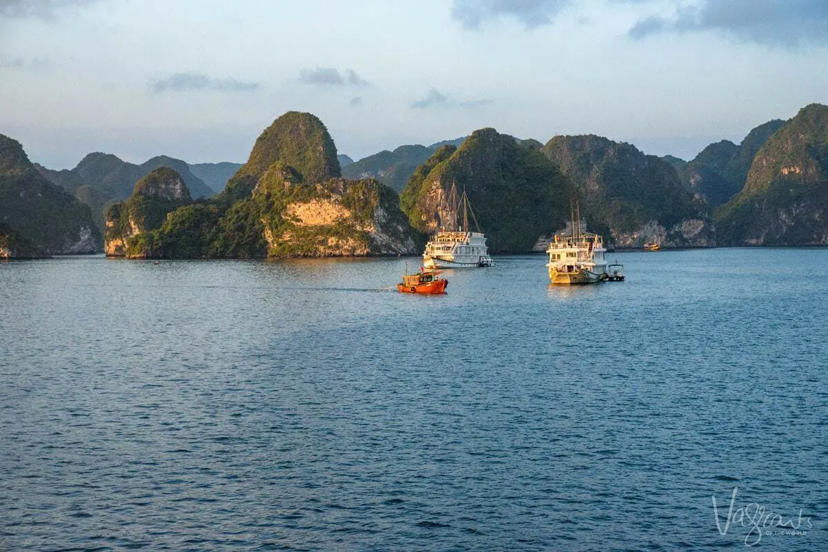 A red fishing boat cruises past the luxury Halong Bay boat cruises as the sun rises. 