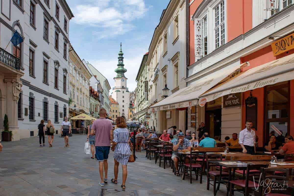 People walking down a shopping esplanade with a church steeple in the background and diners eating alfresco along the way.