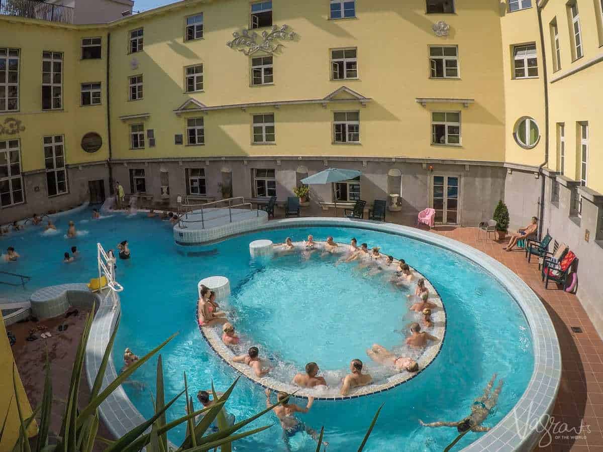 People sitting in the thermal baths in Budapest. A horse shoe shaped sitting area in the pool with the old baths building in the background. 