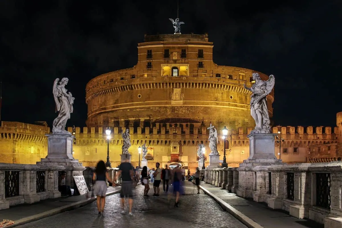 Golden glow of Castel Sant’Angelo at night with people standing in front. 