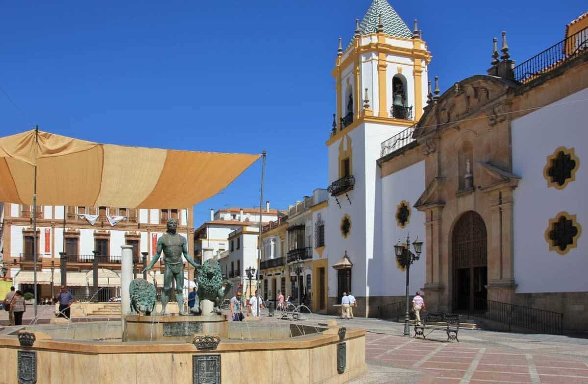 Brass man statue in fountain in square surrounded by white and gold church. Base yourself in Seville and add to you Seville itinerary Day trips from Seville to Ronda