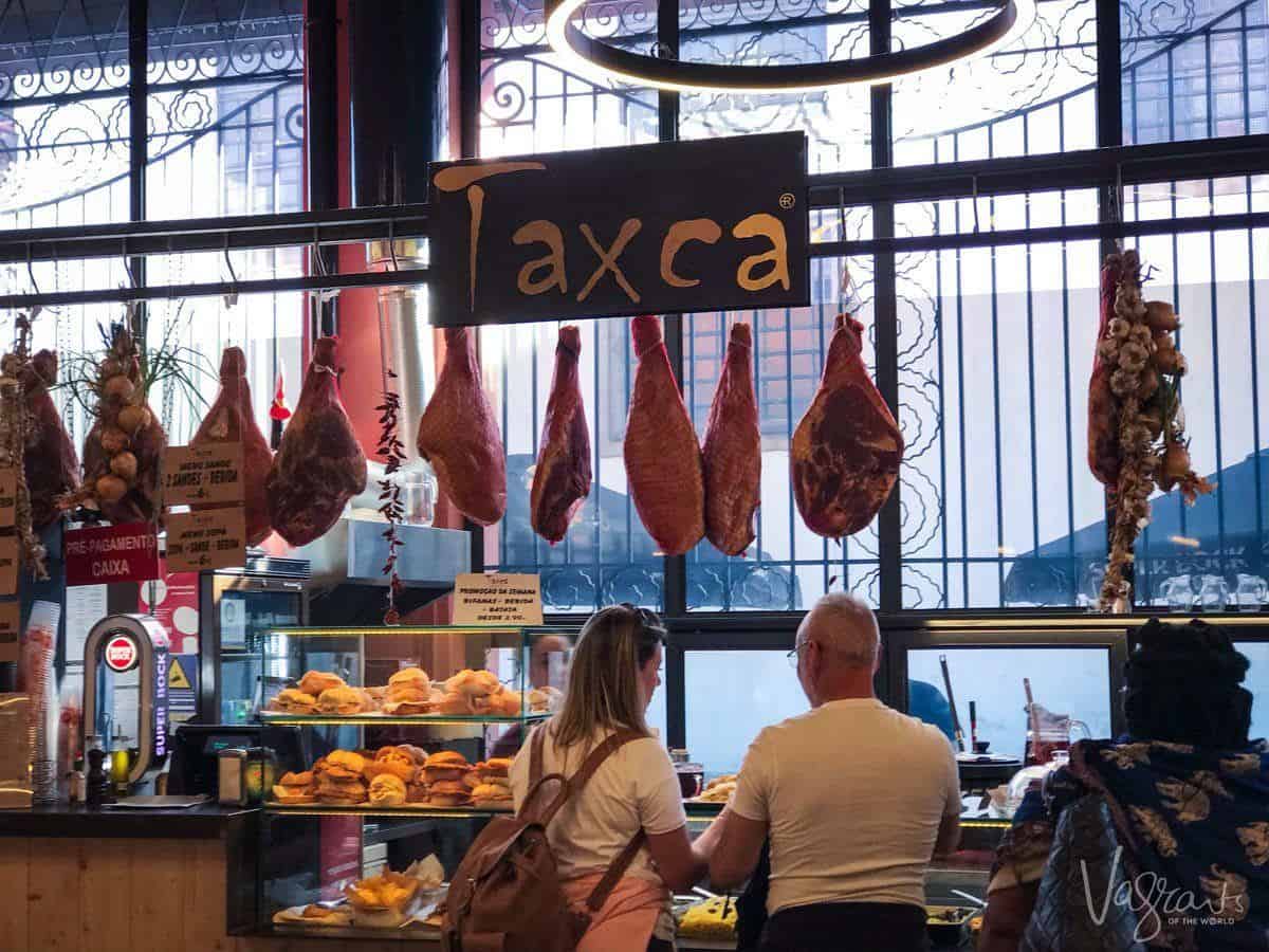 Two people buying lunch at Mercado Beira-Rio with the ham legs hanging from a rack above them. 