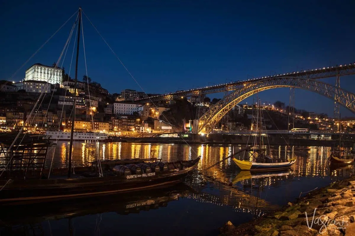 A night view of the Douro river and the illuminated D. Luis I bridge with a port boat in the foreground. 