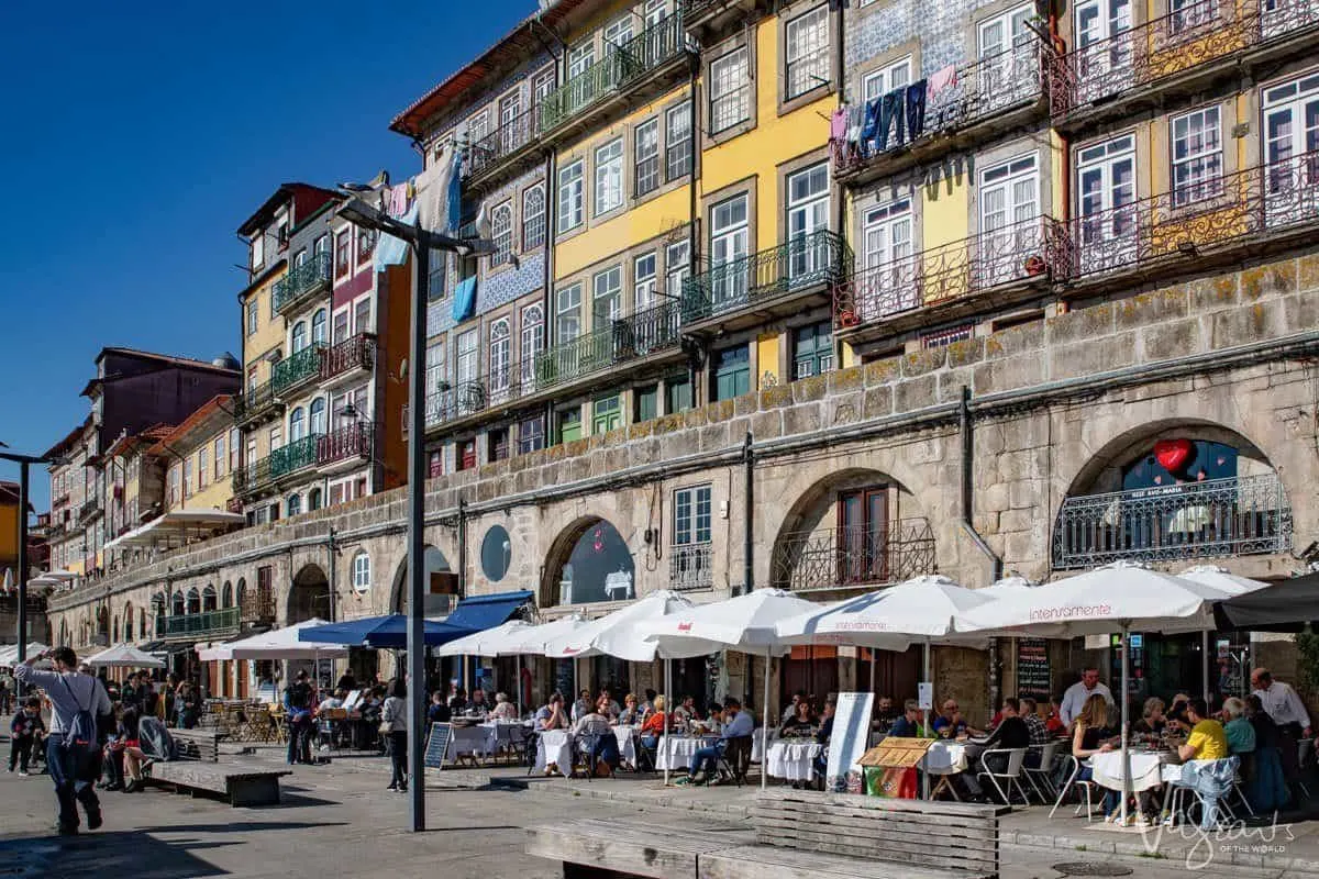 Al fresco dining in Porto under umbrellas beside yellow and green skinny buildings with intricate tile patterns on some of the walls. 