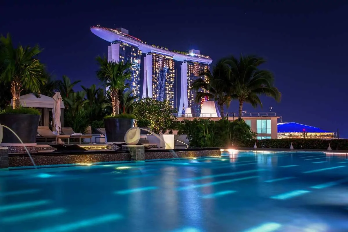 Fountains into a swimming pool with purple lit hotel in background. 