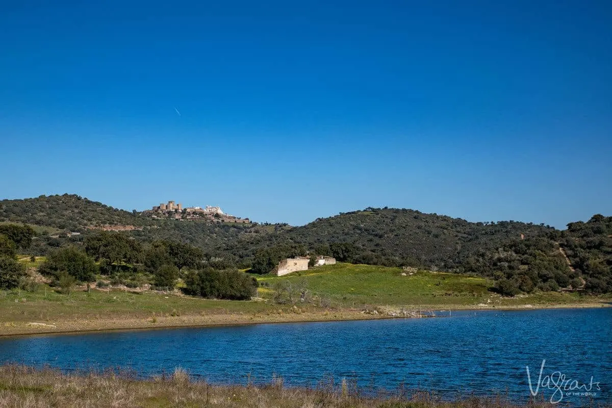 the blue waters of Alqueva lake with hillside village in the background. Stop here for a lunch of alentejo food and wine on your alentejo portugal road trip