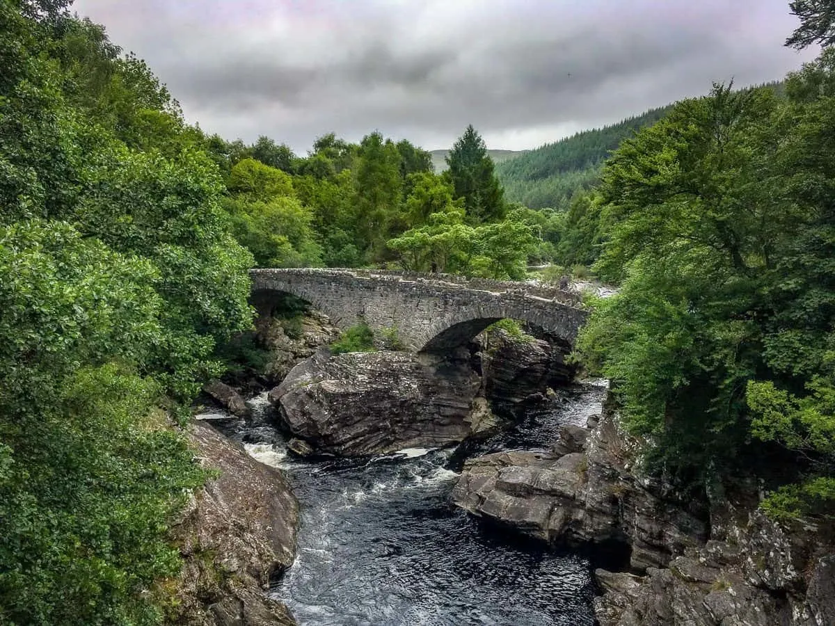 stream flowing under old stone bridge bordered with evergreen trees under a cloudy sky.  the best way to travel on foot is by organising a self guided walking holiday 