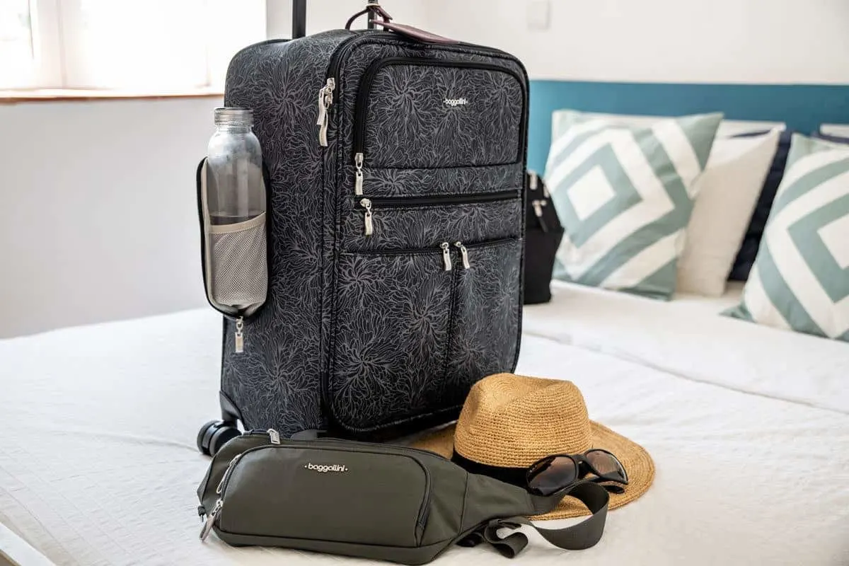 grey baggallini travel bag on a bed with a bum bag and hat.  if you are looking for the  best luggage for travel then you cant beat these travel bags for women
