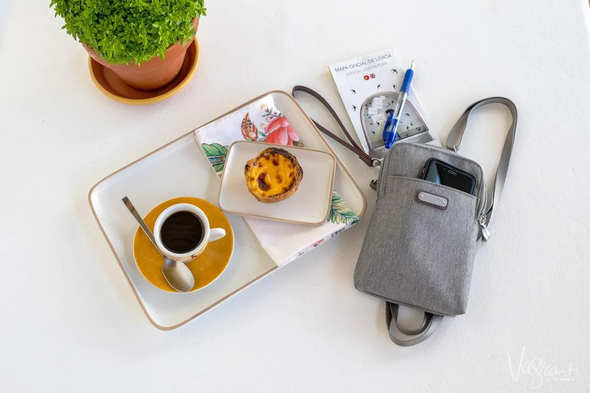 a womens cross body travel bag on a table next to a cup of coffee and a pastel de nata.  this may be the best travel purse for you