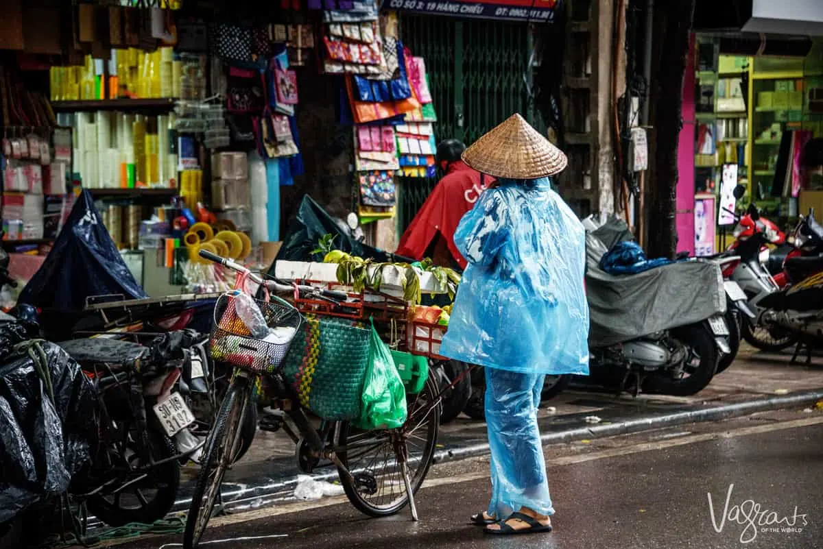 Lady in 2 piece blue rain coat with conical hat next to her bicycle. Watching the locals get around is one of the best free things to do in Hanoi Vietnam.