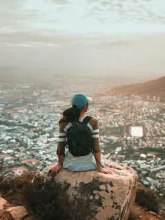Female traveller wearing a backpack sitting on a rock at a lookout over a city and harbour.