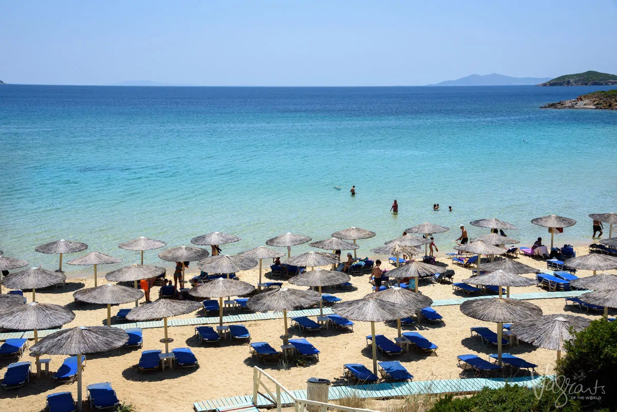 A beach full of sun beds and shades leading down to blue clear water in Greece.