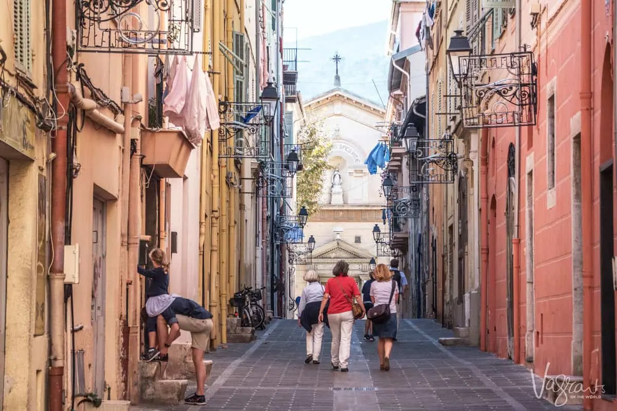 Tourists walking down a narrow street in France while a man ties his shoe. 