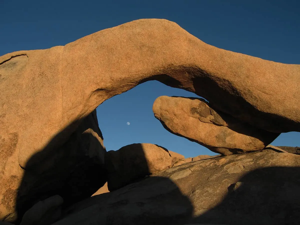 Arch Rock against a dark sky with moon. Arch Rock trail is one of the most popular hikes in Joshua Tree