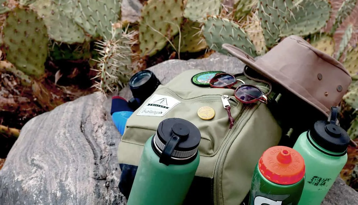Backpack, hat, sunglasses and water bottles lying on rocks among cacti. 