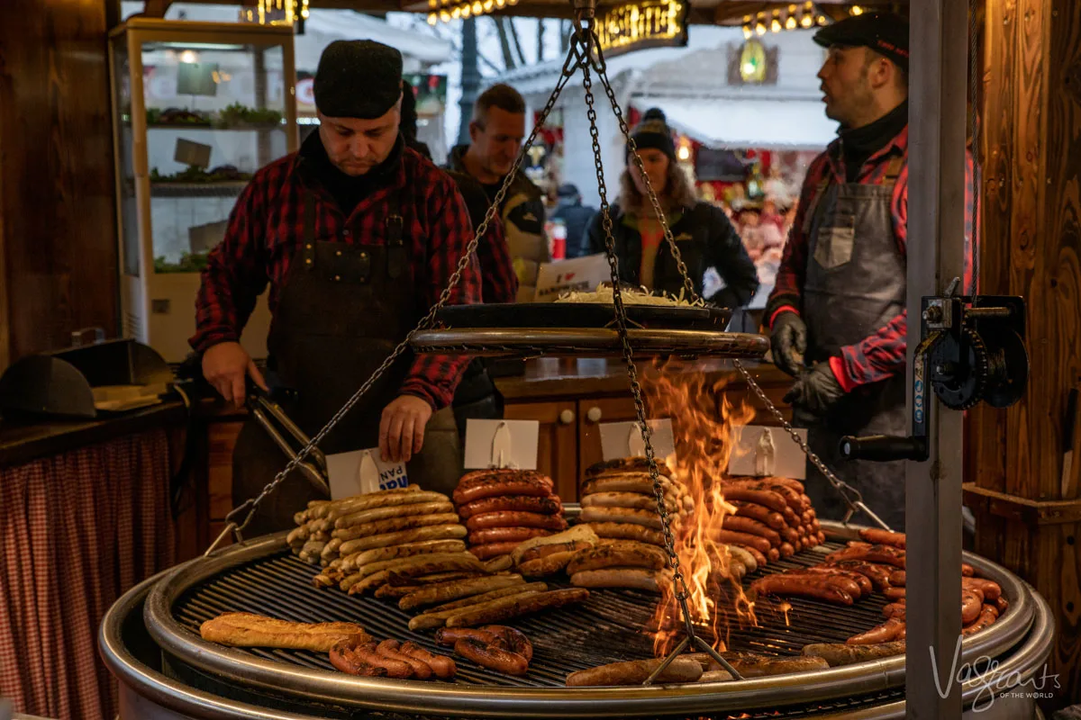 Men prepare large quantities of assorted sausages over an open flame on a circular, swinging grill. A typical sight at Europe's best Christmas markets.