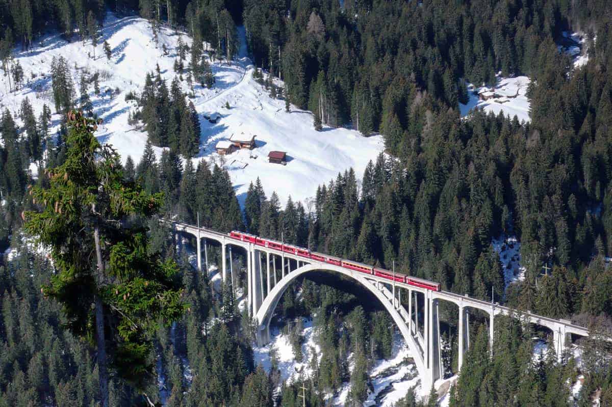 Red train passing over suspension bridge above snow covered forest.