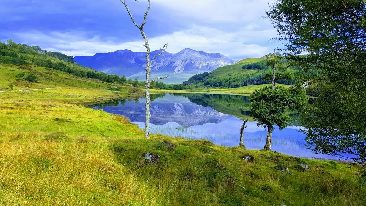  Beinn Eighe, Scotland with lush green grass and snow dusted mountain reflections in the river. Hiking in Scotland, or Hill Walking as it is known, offers hikers beautiful landscapes like these.