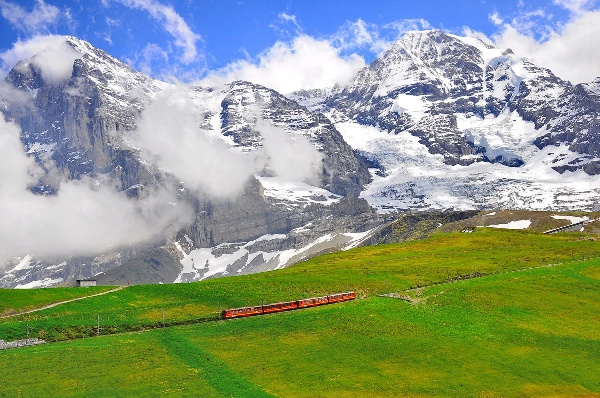One of Switzerland's biggest attractions is the Jungfraujoch Train crossing a lush green field with the snow covered Swiss Alps in the background. 