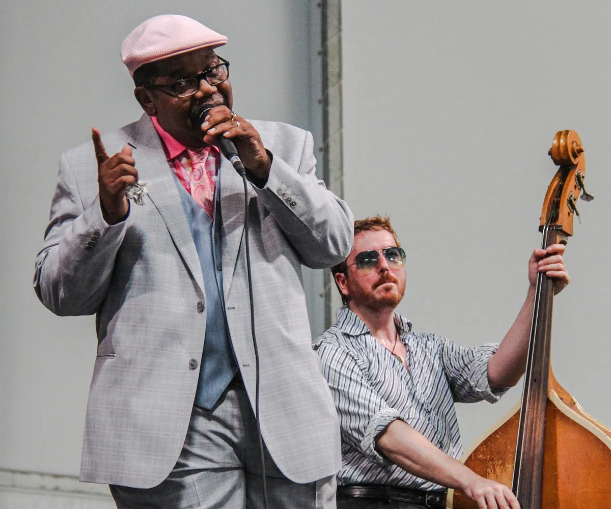 Black man in a suit and hat sinnging jazz with a bass player sitting behind at the Satchmo summerfest in New Orleans