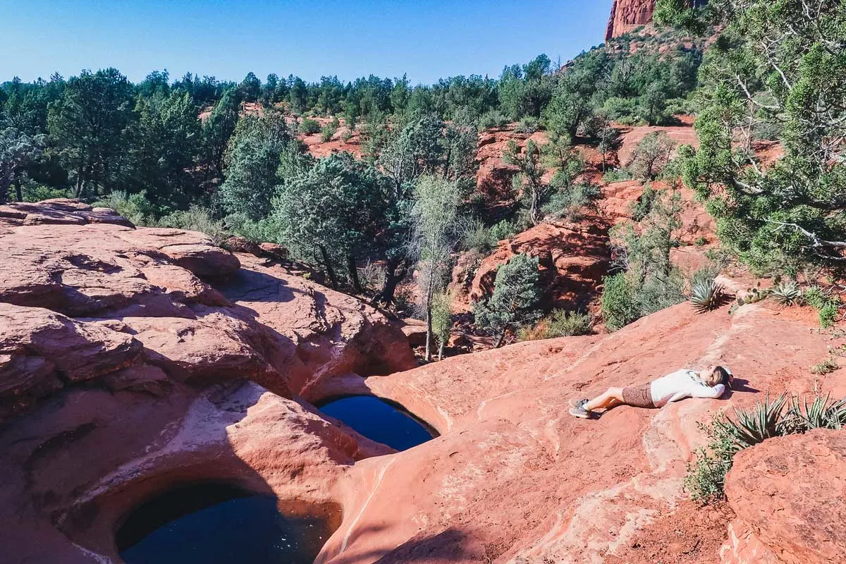 Hiker laying in the sun on red rocks next to 2 natural pools at 7 sacred pools. 