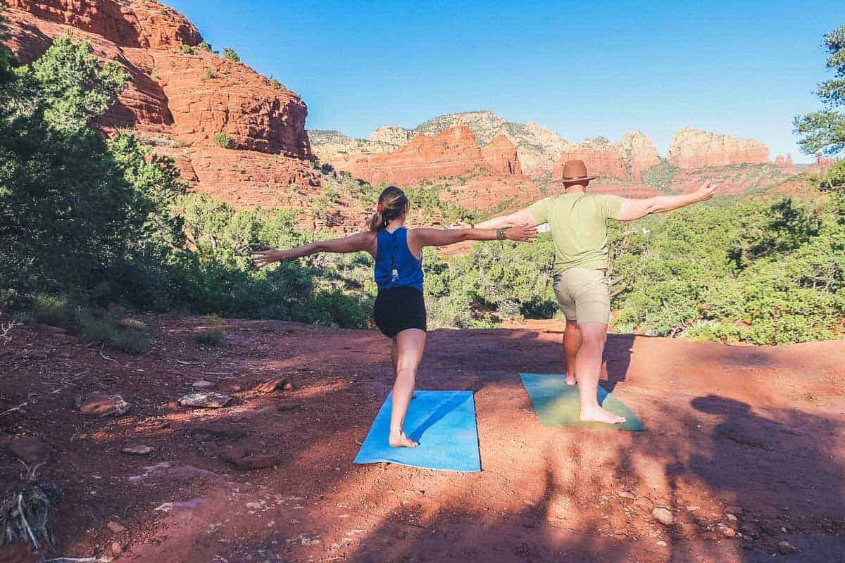 A couple standing on a red rocky outcrop with arms stretch in Vortex Yoga pose. 
