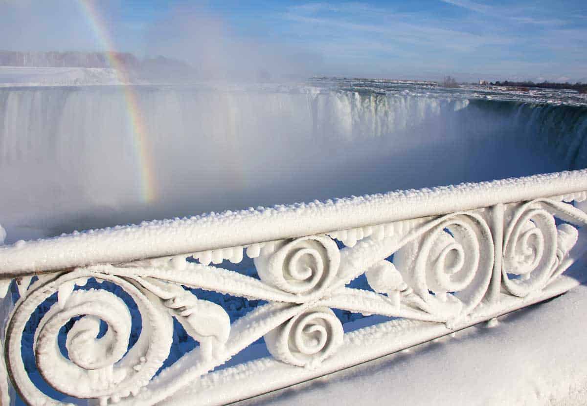 Ice covered ornate railing overlooking a rainbow across Niagara Falls.