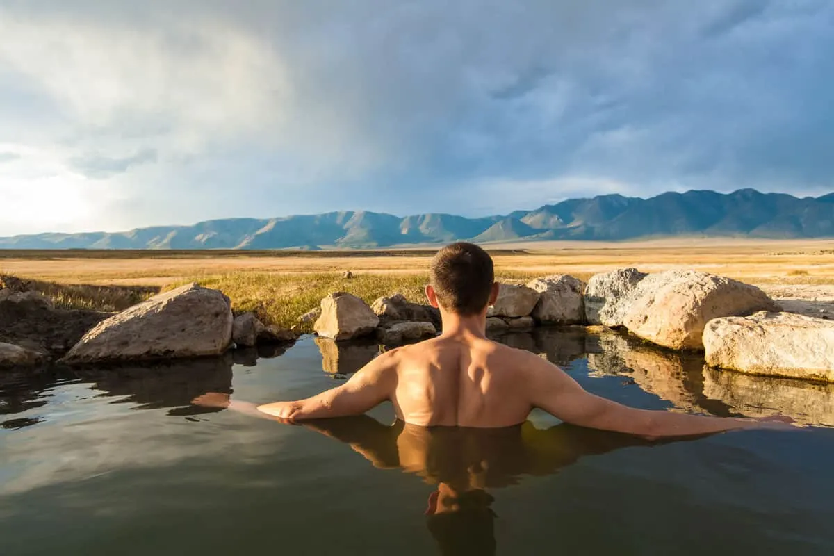 Man sitting in a natural hot spring in a field with mountains in the distance. 