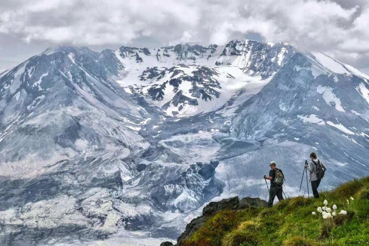 COuple trekking around Mount ST. Helen on a grassy outcrop against the white volcanic mountain.