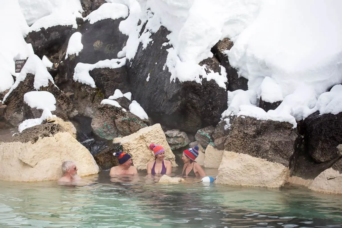 People sitting in thermal hot springs wearing winter beanies with snow in background.